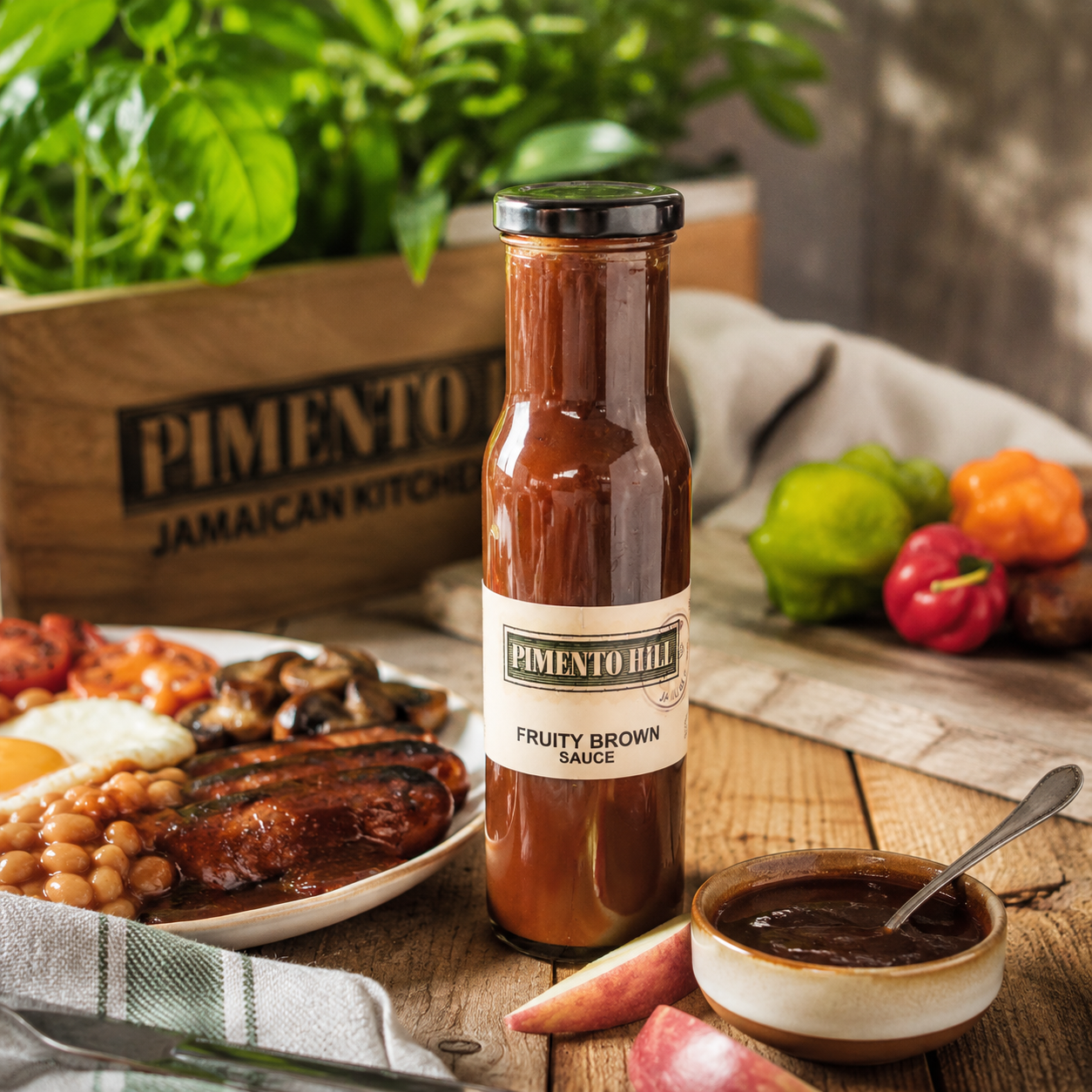 Pimento Hill Fruity Brown Sauce styled on a rustic wooden table alongside a cooked breakfast, apples and Caribbean-inspired ingredients in a warm kitchen setting.